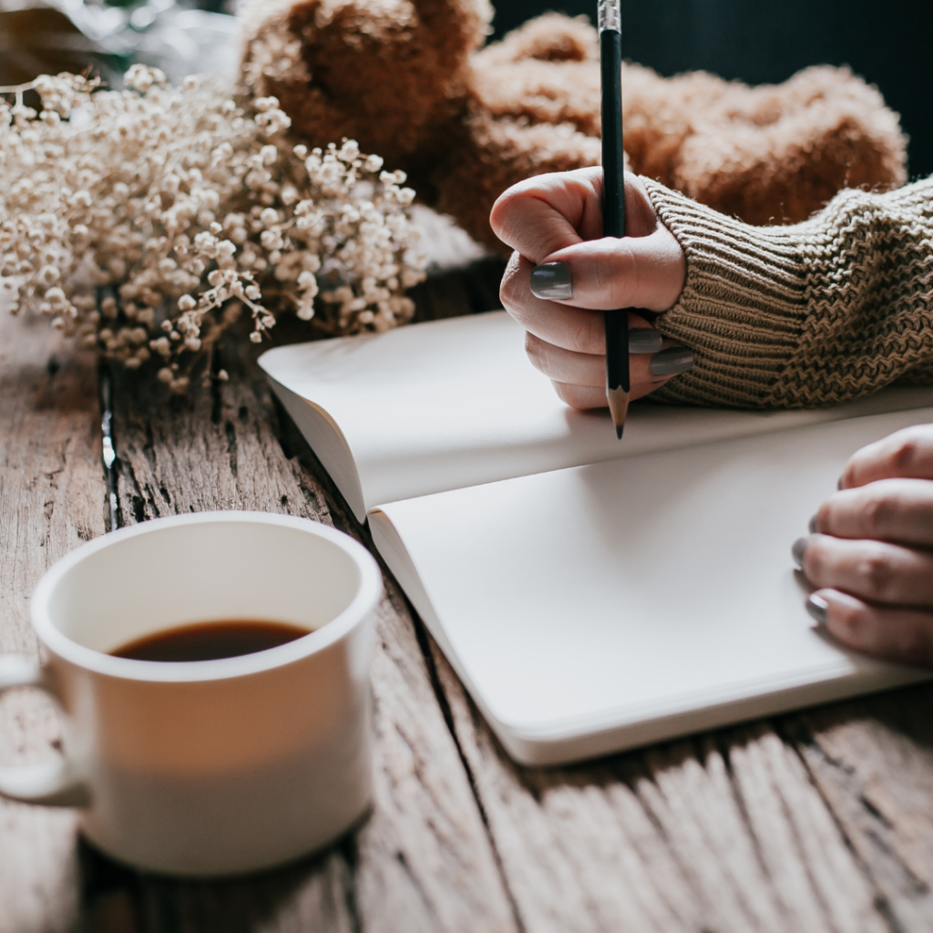 book on wood table with hand and pencil over top, coffee mug and dried flowers in back - canva pro image