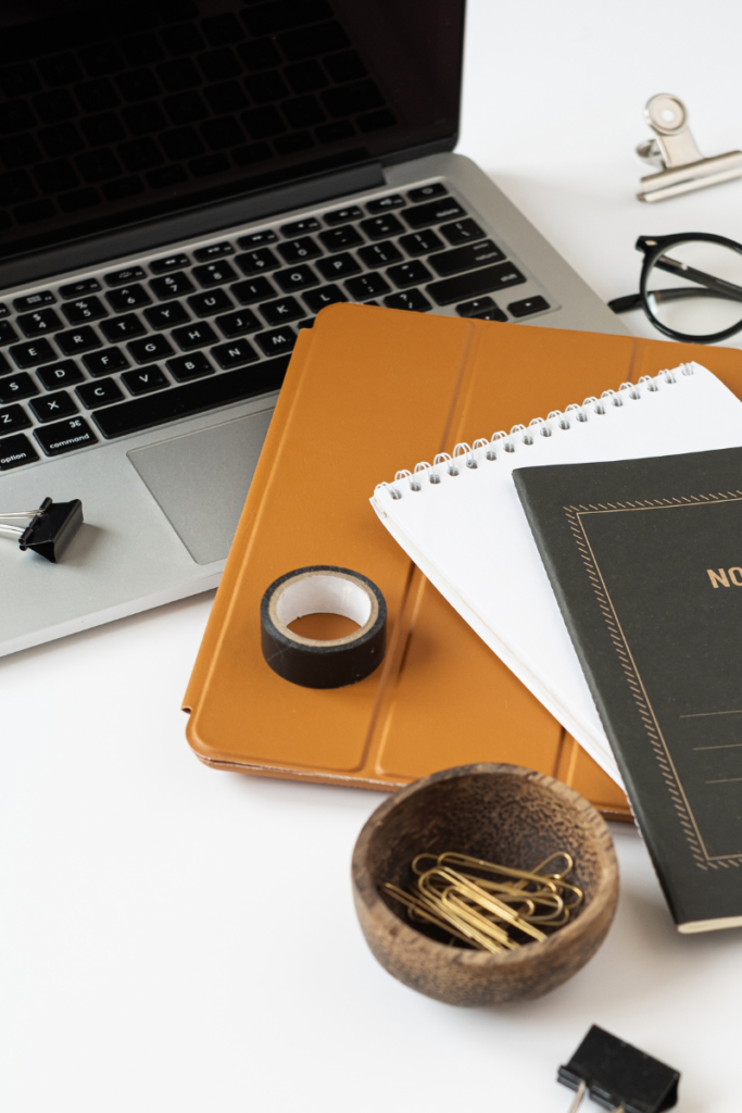 Photo of laptop, ipad in a brown case, some paperclips and note pad