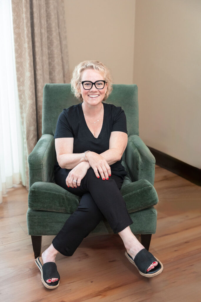 cindy gordon dressed in all black, sitting in a green chair with a brown floor, yellow walls and pale yellow curtains behind her.