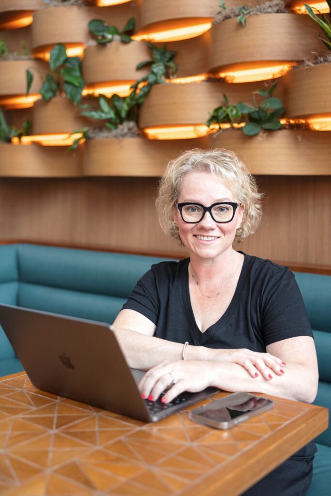 Cindy gordon sitting at a brown table, dressed in all black with her computer, she is smiling at the camera during a business coaching session