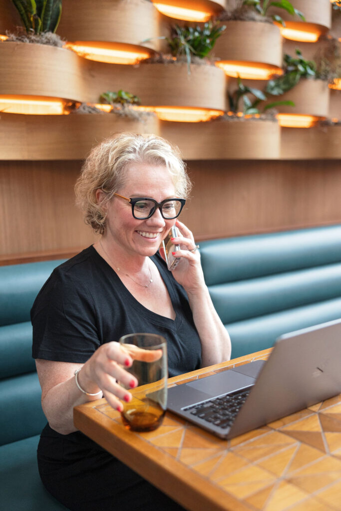 Cindy Gordon dressed in all black sitting in a restaurant, looking at her computer, talking on the phone to a business coaching client and has her hand on a water glass.