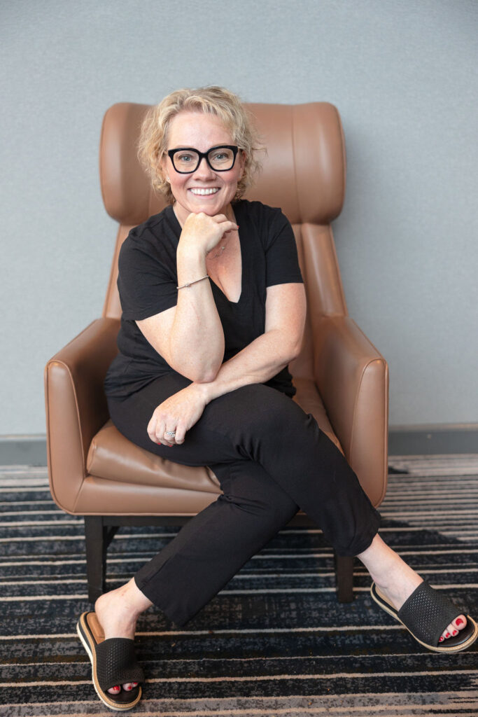 picture of cindy gordin dressed in a black shirt and black pants sitting cross legged on a brown chair. Her hand is holding up a chin, she is smiling before a business coaching session