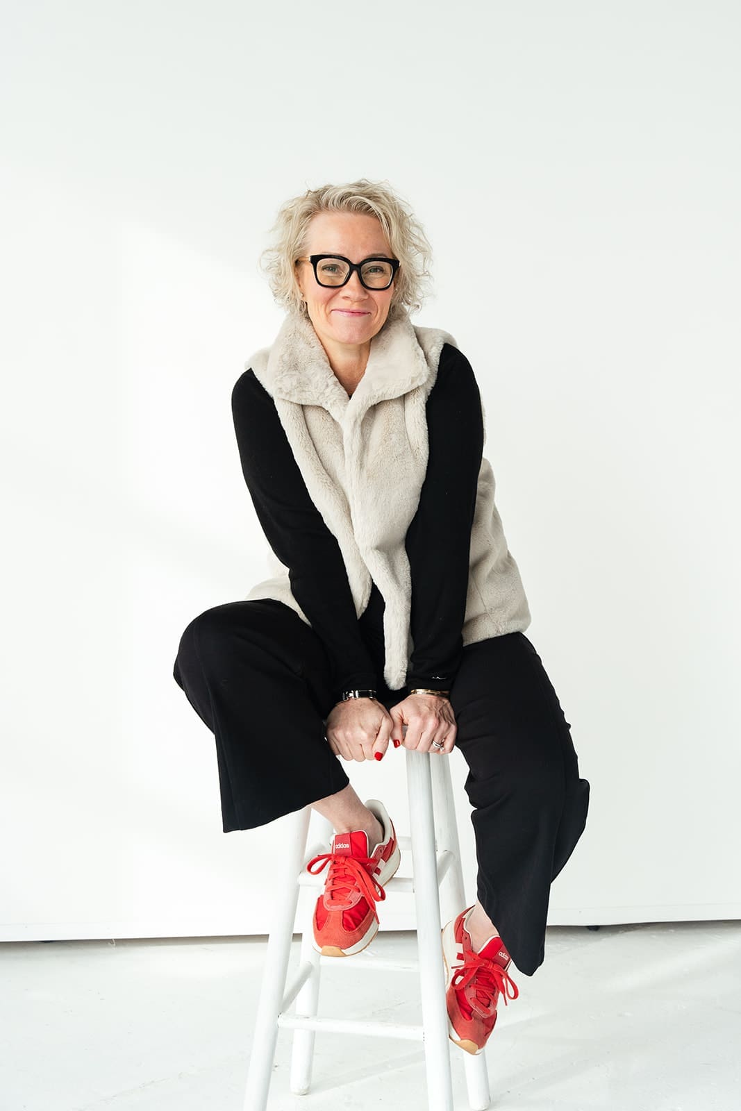 Cindy Gordon seated on a white stool in a cream vest, black trousers and red shoes