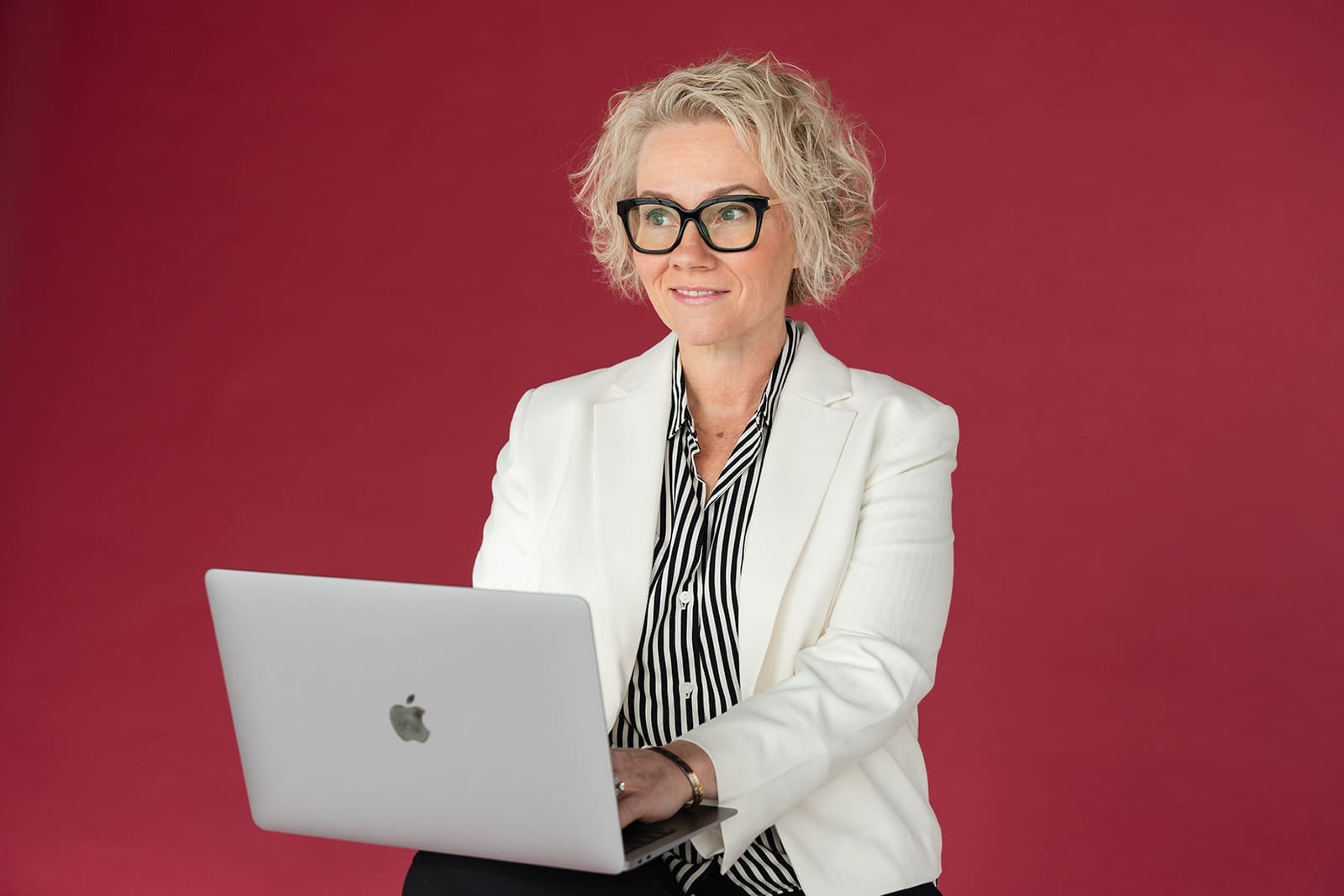 Cindy Gordon holding an open silver laptop in a white blazer against a deep red background